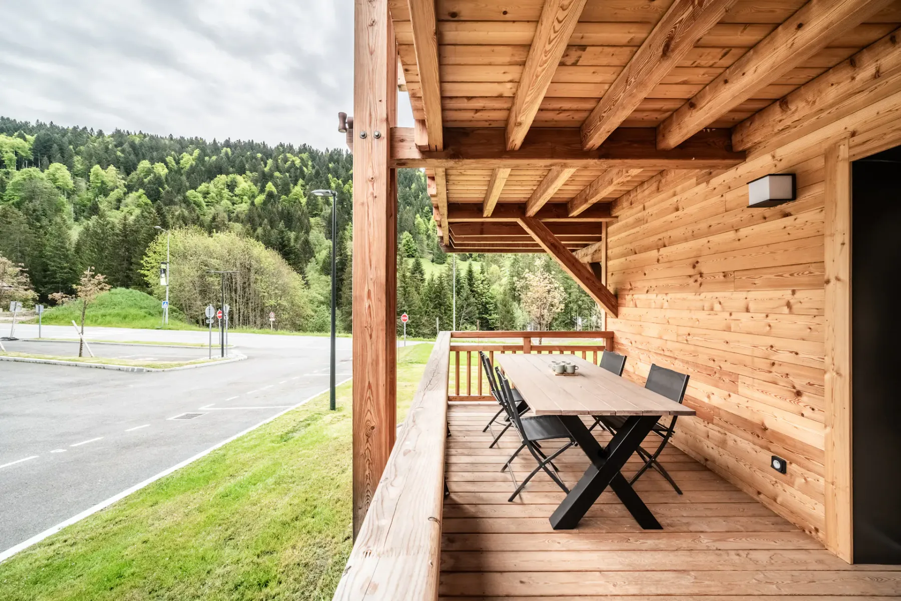 Un patio de madera cubierto tiene una mesa de comedor y sillas junto a una carretera arbolada.