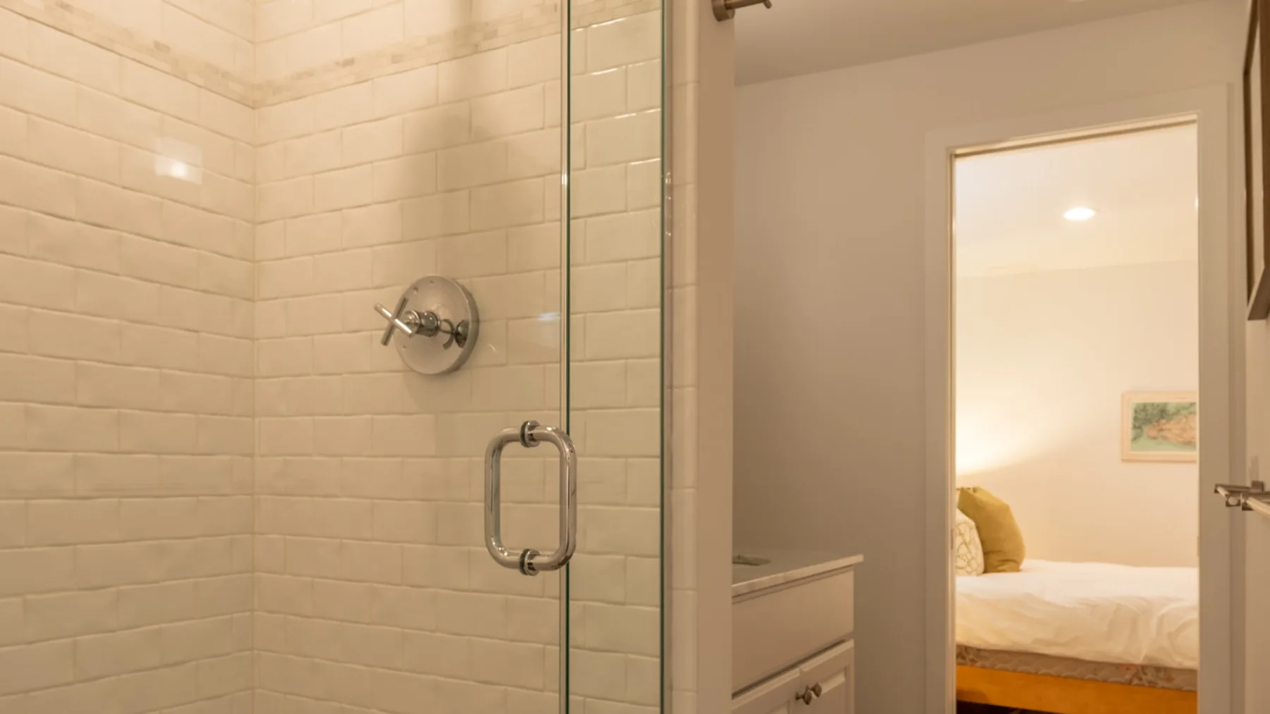 Un baño con azulejos y ducha de vidrio junto a una puerta abierta hacia un dormitorio.