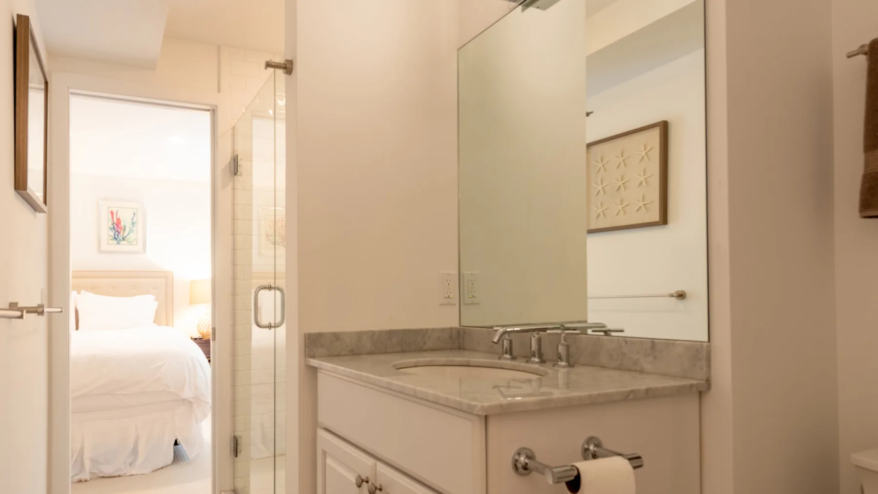 Un lavabo de baño con espejo junto a una ducha de vidrio, con un dormitorio visible por la puerta.