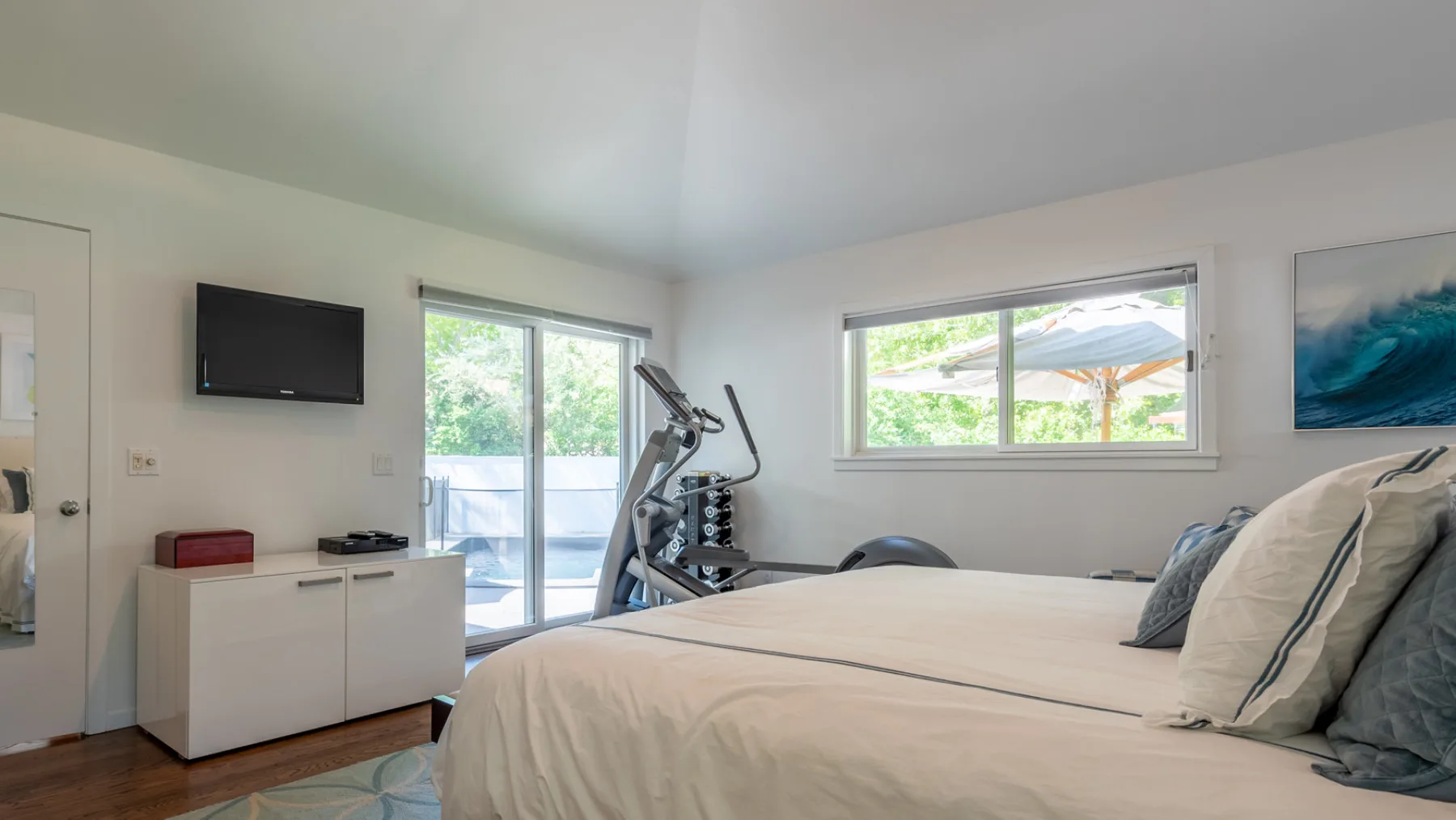 Un dormitorio luminoso con una cama, equipo de ejercicio y ventanas con vistas a la vegetación.