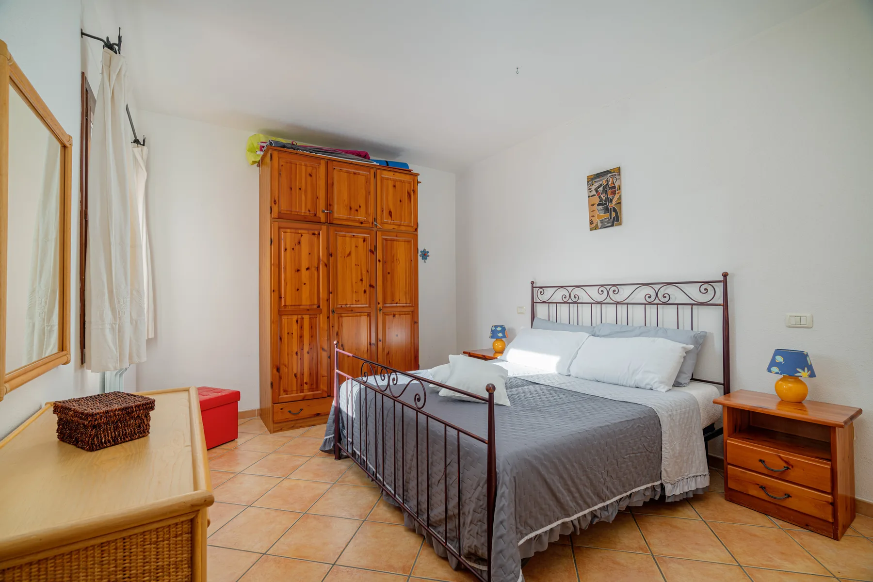 Un dormitorio con una cama de estructura metálica, un armario de madera y suelo de baldosas.
