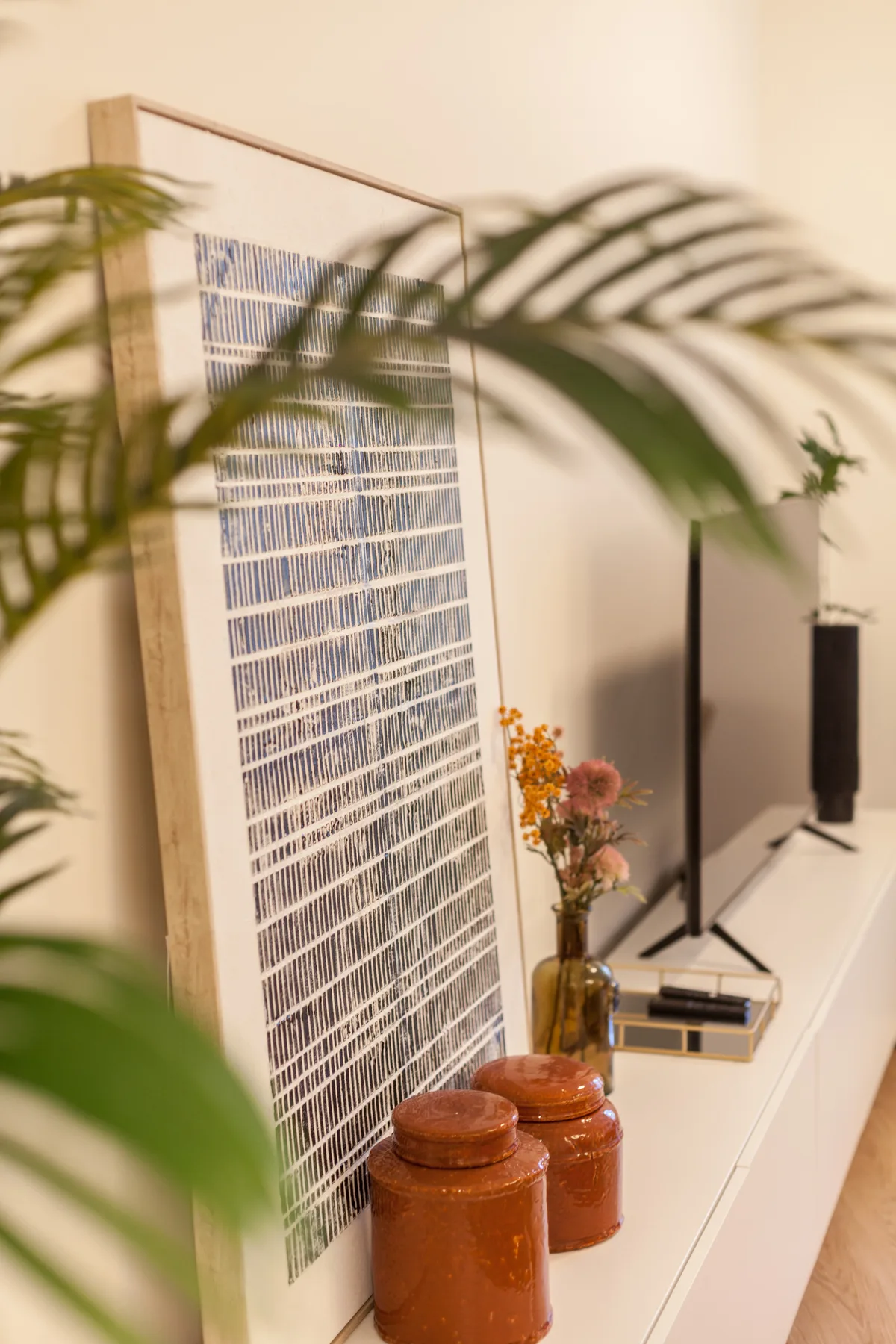 Un rincón de sala muestra una obra enmarcada, recipientes decorativos y flores junto a un televisor.