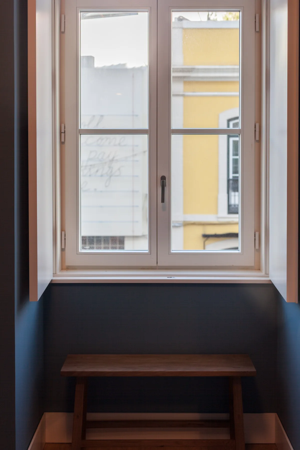 Una ventana sobre un banco de madera, con un edificio amarillo visible afuera.