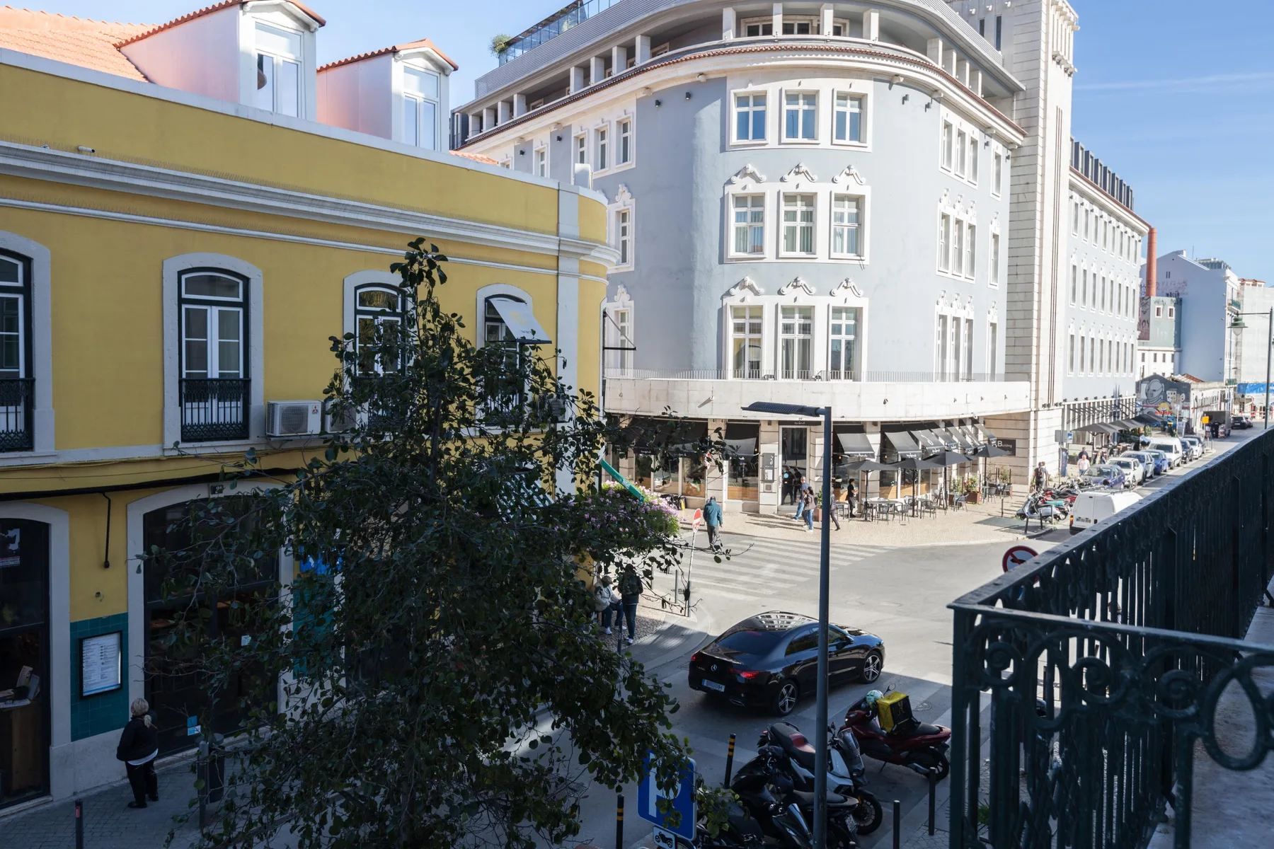 Escena urbana con edificios coloridos, mesas de café al aire libre y vehículos estacionados.