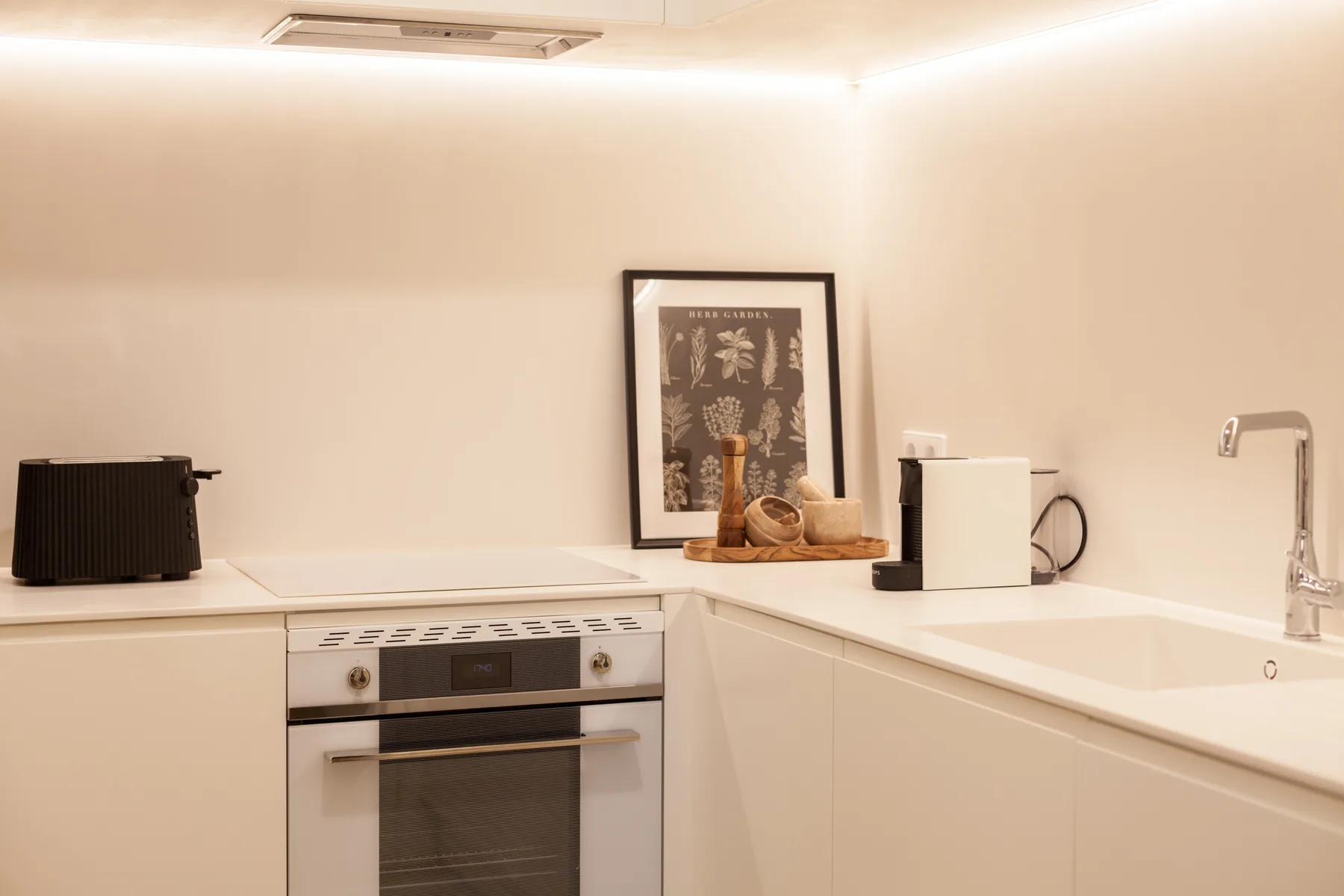 Una cocina moderna y luminosa con encimeras blancas, horno y una lámina enmarcada.