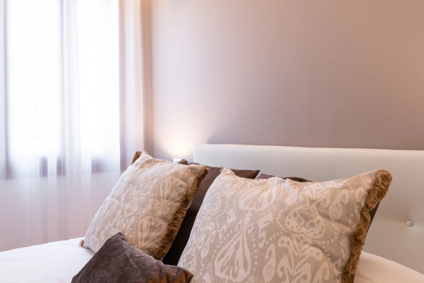 Un dormitorio con cojines estampados sobre una cama junto a una ventana luminosa.