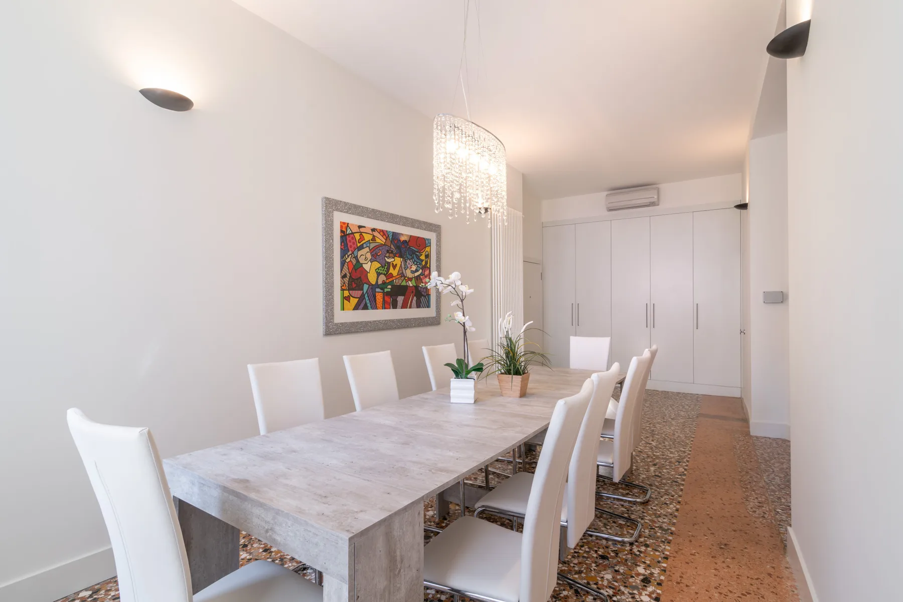 Un comedor luminoso con una mesa larga, asientos blancos y arte colorido en la pared.