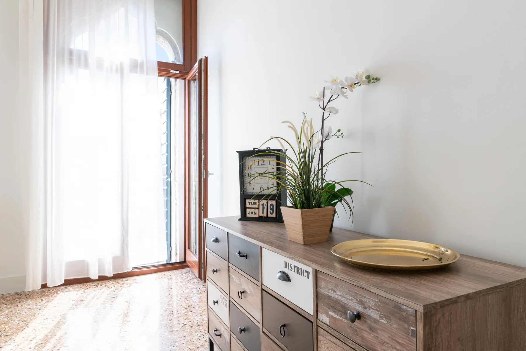 Una habitación luminosa con una cómoda de madera, una planta en maceta y cortinas translúcidas junto a la ventana.