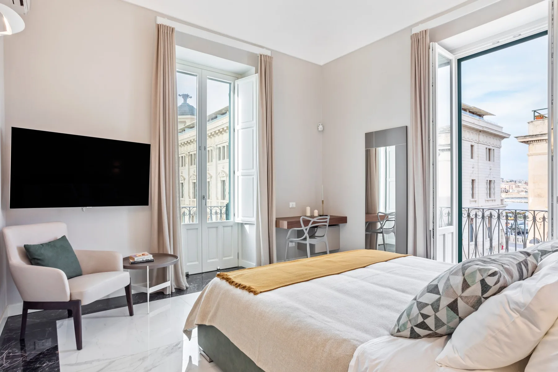 A bedroom with a large bed, a wall-mounted television, and tall windows opening onto a balcony.