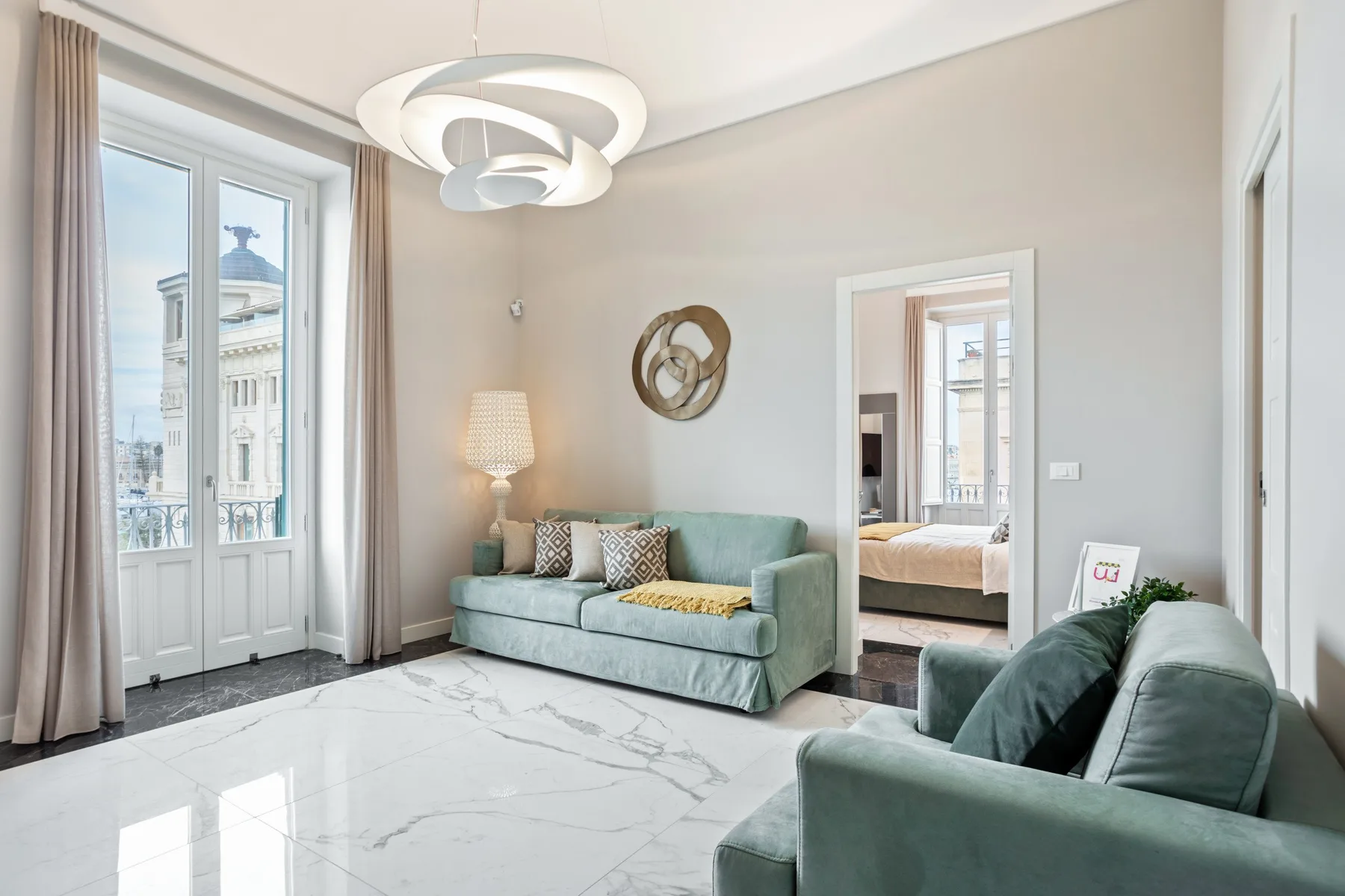 A bright living room with green seating, a large window, and a view into a bedroom.