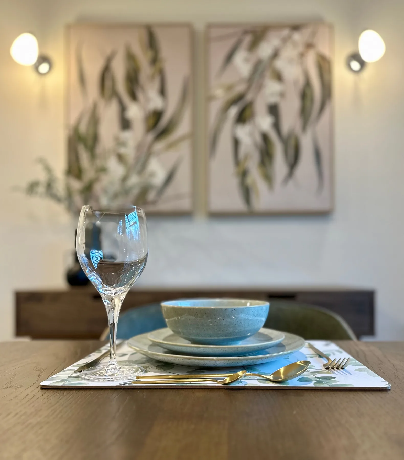 Una mesa de comedor está preparada con plato, cuenco y copa bajo cuadros botánicos.