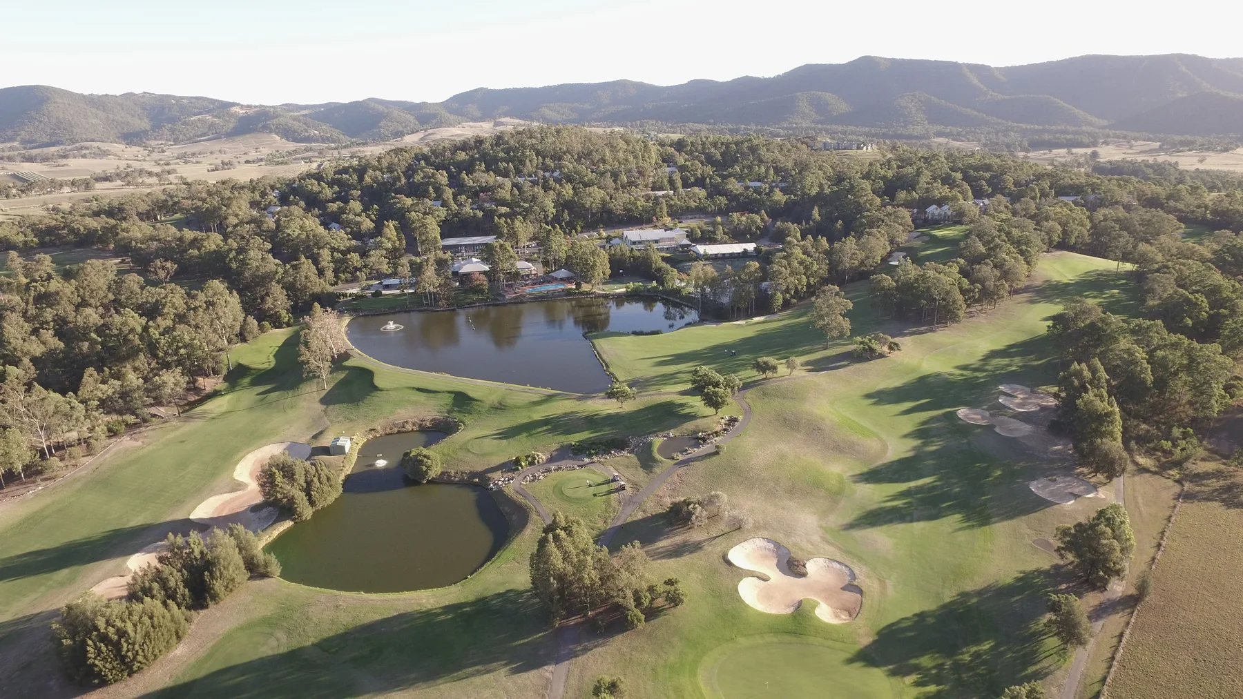 Vista aérea de un campo de golf con estanques y colinas arboladas al fondo.