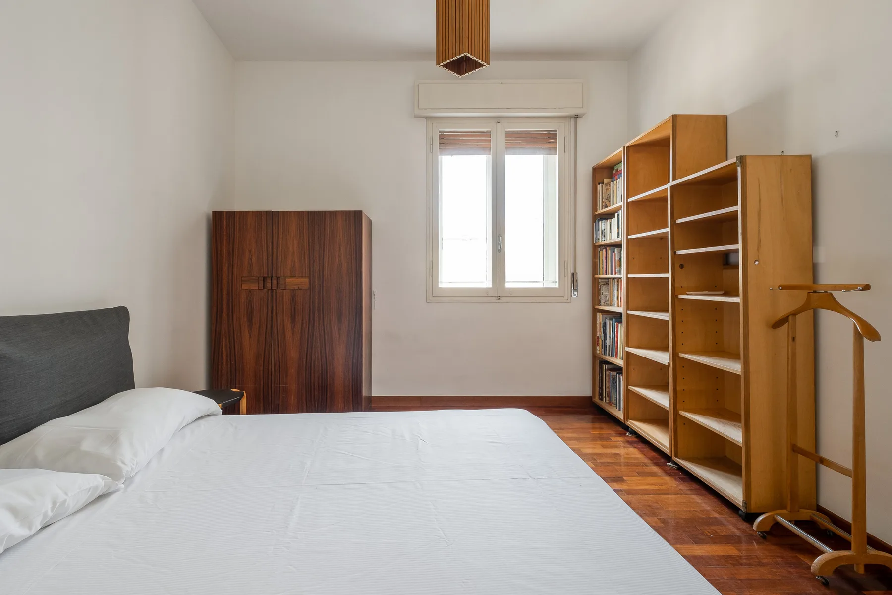 A bedroom with a bed, a wardrobe, and wooden shelving beside a window.