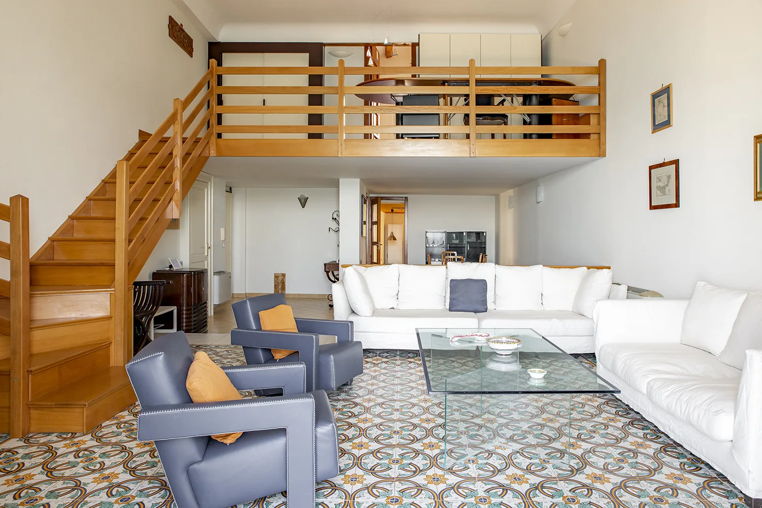 Una sala de estar con asientos blancos, sillones azules y una escalera de madera hacia un altillo.