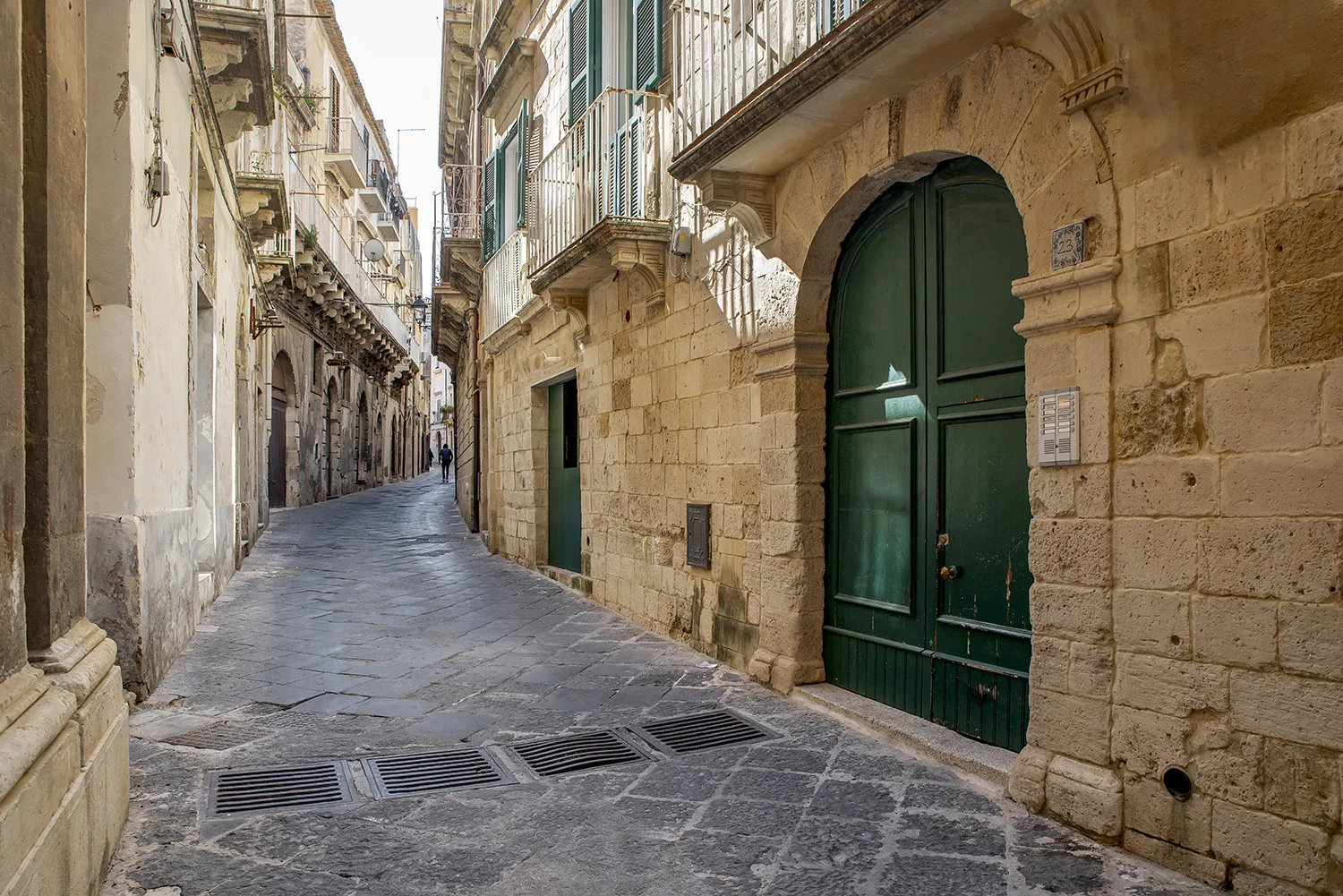 Una calle estrecha pavimentada con piedra pasa entre edificios cercanos con puertas y contraventanas verdes.