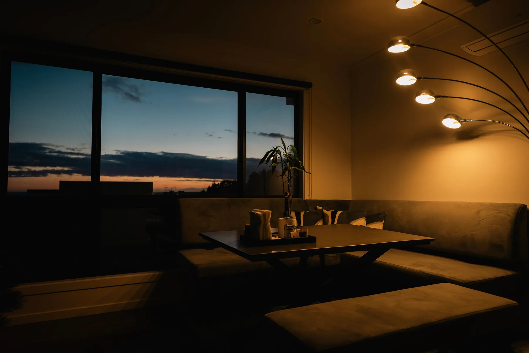 Una sala con poca luz tiene asientos bajos y una gran ventana con vistas al cielo del atardecer.