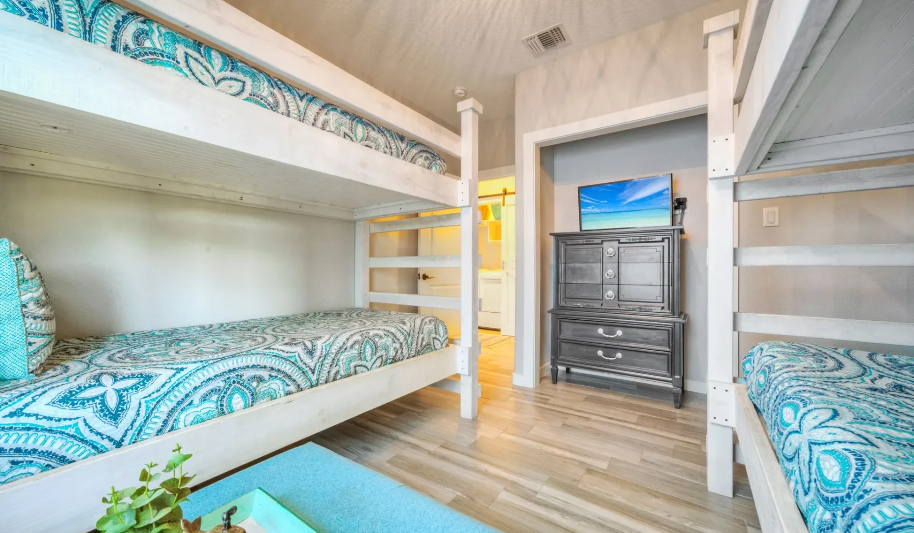 Un dormitorio con literas, ropa de cama turquesa estampada y una cómoda bajo un televisor.