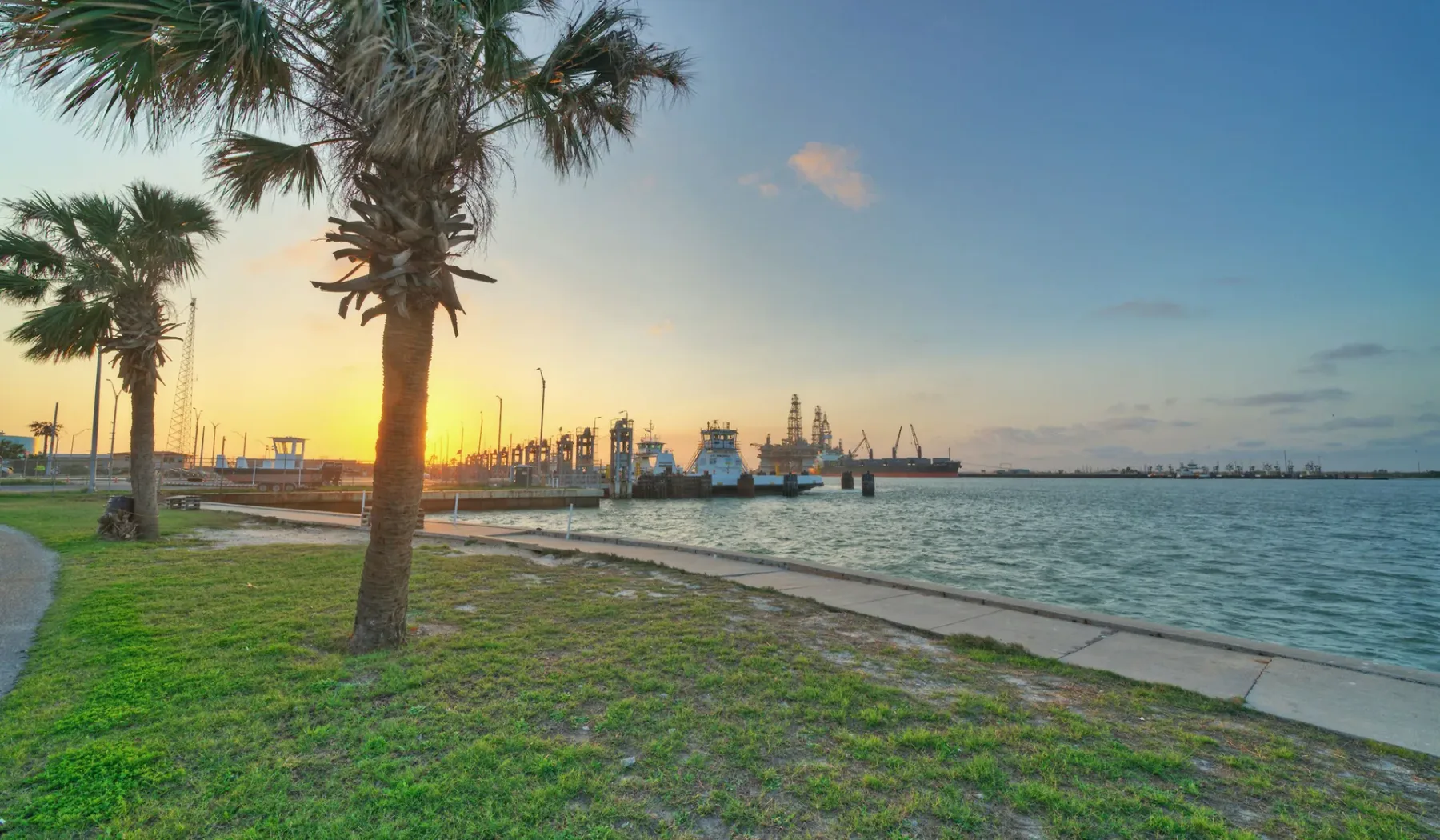 Un paseo junto al agua con palmeras mira hacia barcos al atardecer.