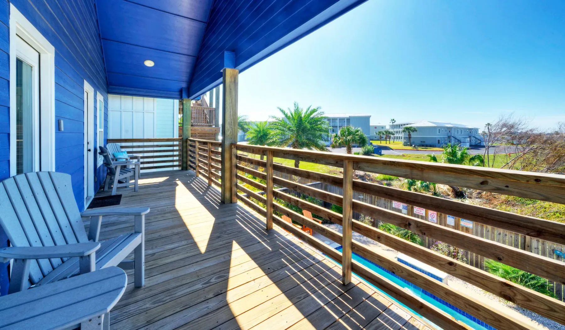 Una terraza de madera cubierta, con revestimiento azul y una barandilla frente a palmeras.