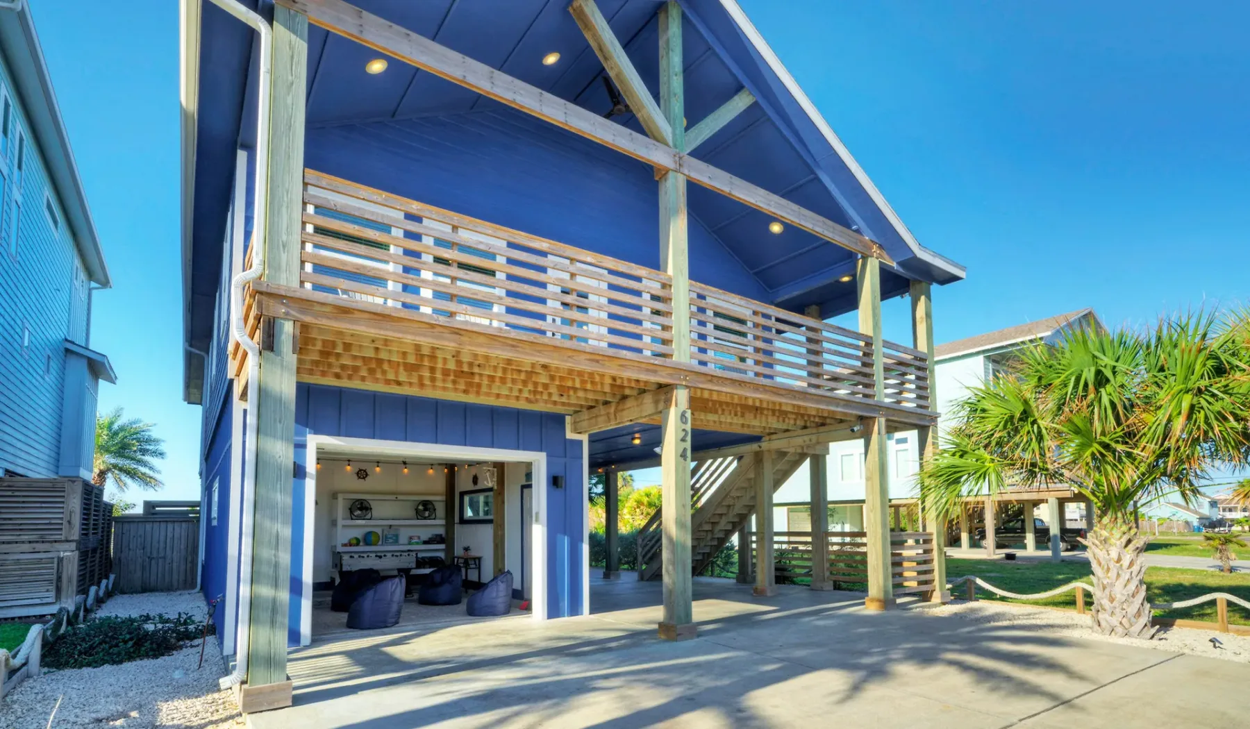 Una casa azul de playa se alza sobre soportes, con una sala abierta bajo el balcón.