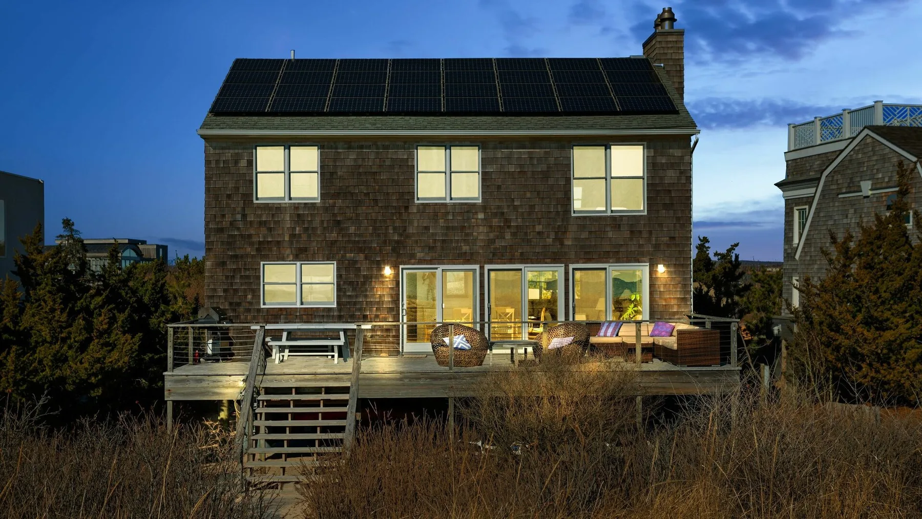 Una casa de dos plantas al anochecer, con paneles solares en el techo y una terraza elevada.