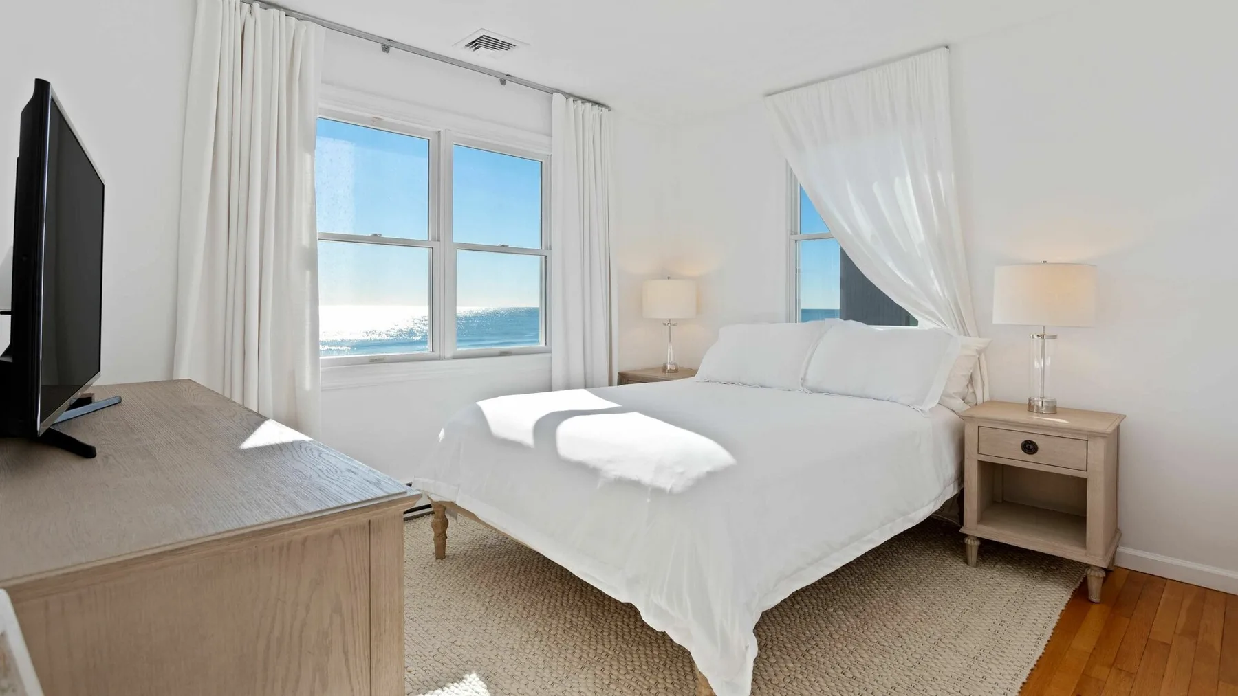 Un dormitorio luminoso con una cama blanca, mesillas de noche y vistas al mar por las ventanas.