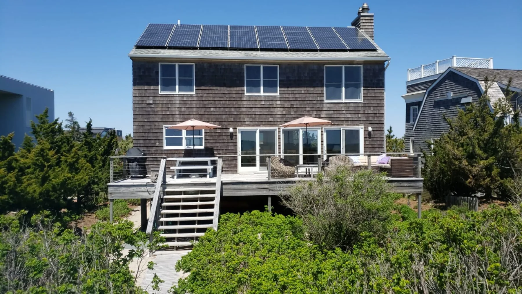 Una casa de tejas oscuras con paneles solares en el techo se alza sobre un jardín frondoso.