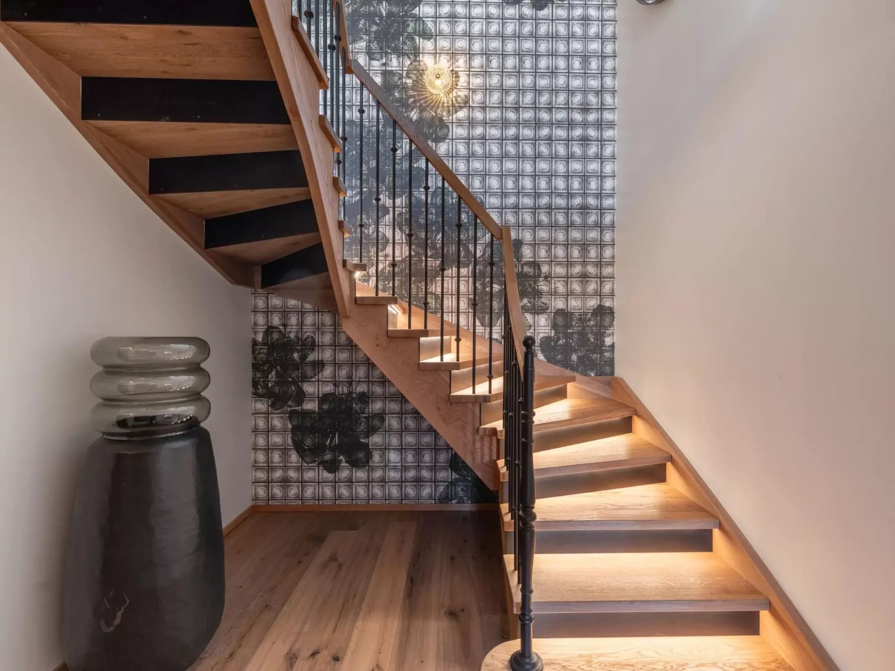 Una escalera de madera sube junto a una pared estampada en un interior con luz cálida.