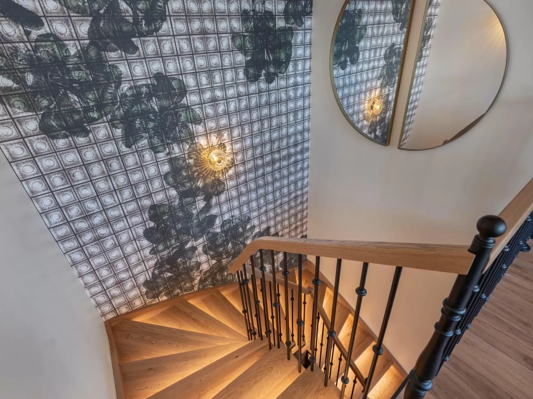 Una escalera curva de madera desciende junto a un papel tapiz estampado y un espejo redondo.