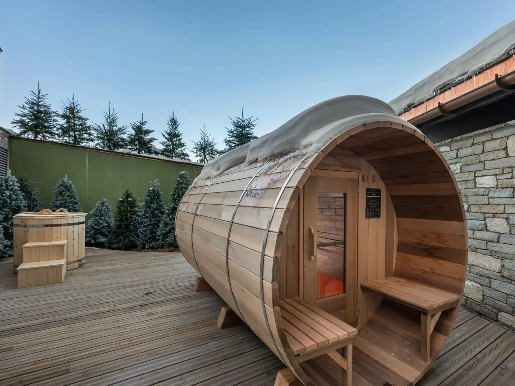Una sauna de madera con forma de barril está sobre una terraza junto a árboles de hoja perenne.