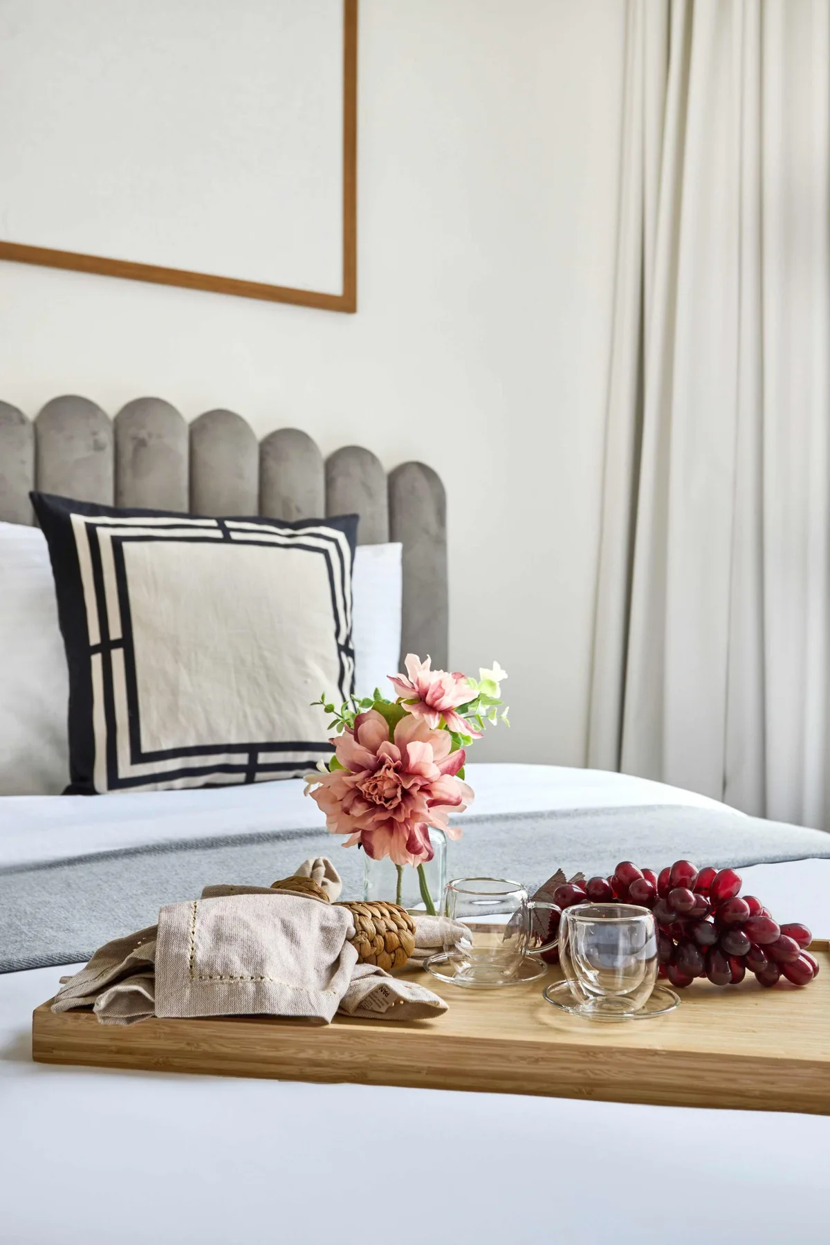 Una cama bien tendida con una bandeja de flores, vasos y uvas en primer plano.