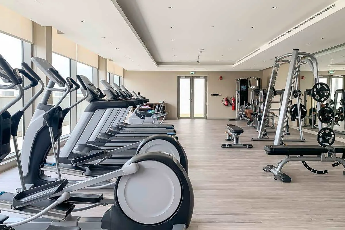 Un gimnasio con máquinas de cardio junto a las ventanas y equipo de pesas.