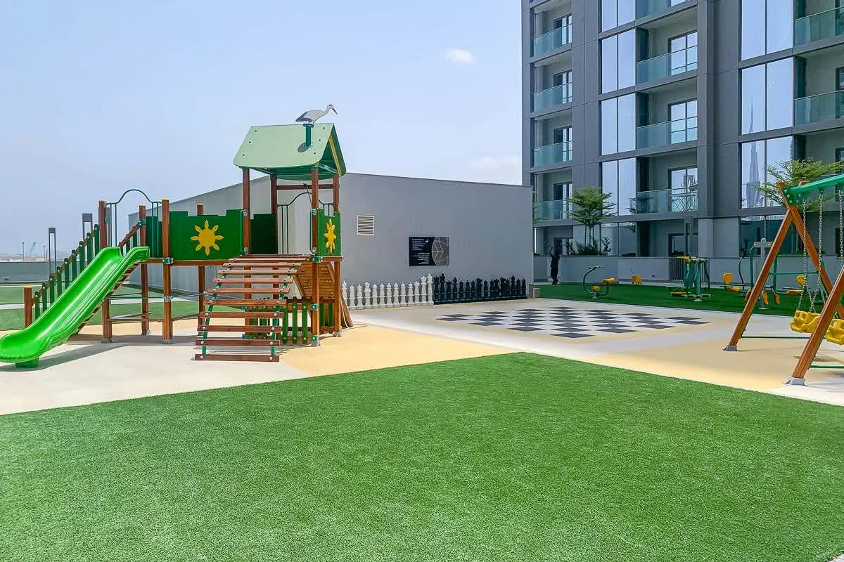 Un parque infantil al aire libre con un tobogán verde y una estructura para trepar junto a un edificio moderno.