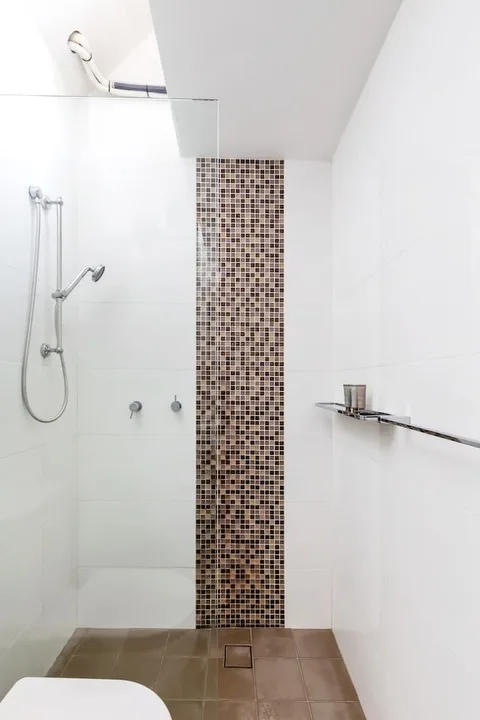 Un baño luminoso con ducha y una estrecha pared decorativa de mosaico.