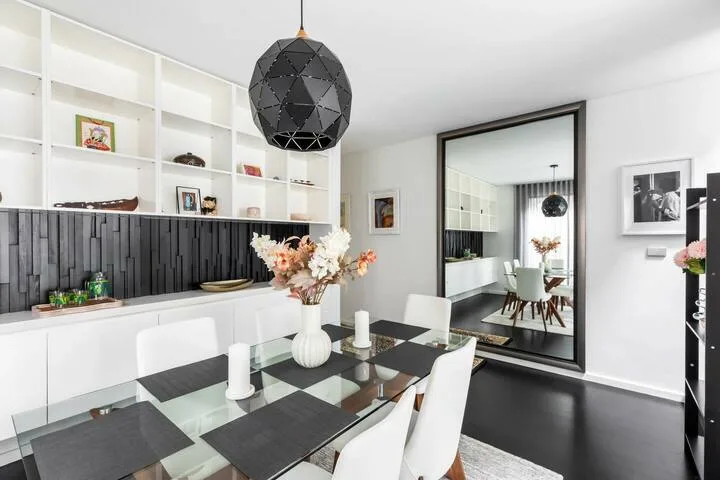 Comedor con mesa de vidrio, asientos blancos y un gran espejo en la pared.