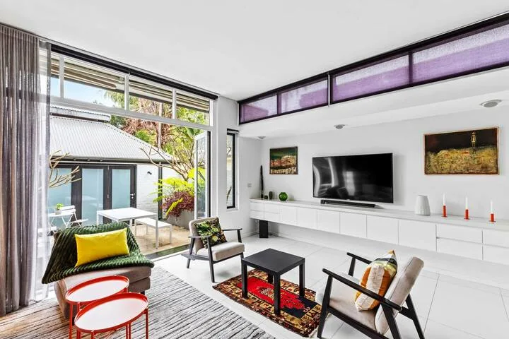 Una luminosa sala de estar con televisor, asientos y grandes ventanas que dan a un patio.