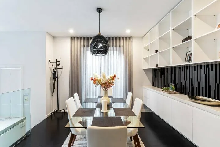 Un comedor luminoso tiene una mesa de vidrio, asientos blancos y ventanas altas.