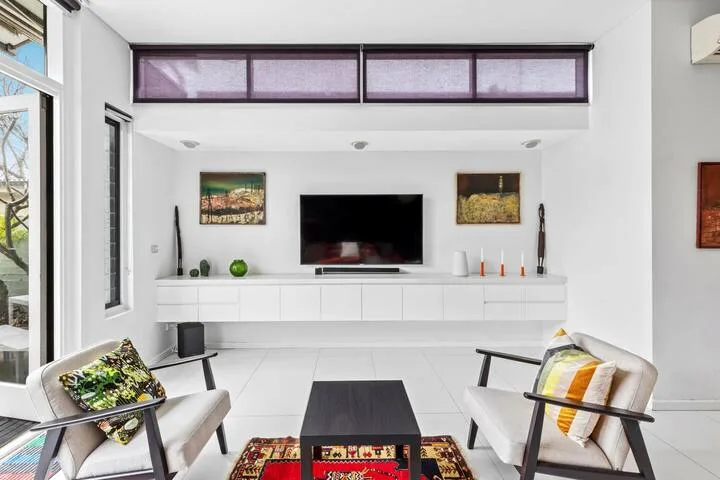 Una sala luminosa tiene un televisor sobre gabinetes blancos y asientos alrededor de una mesa de centro.