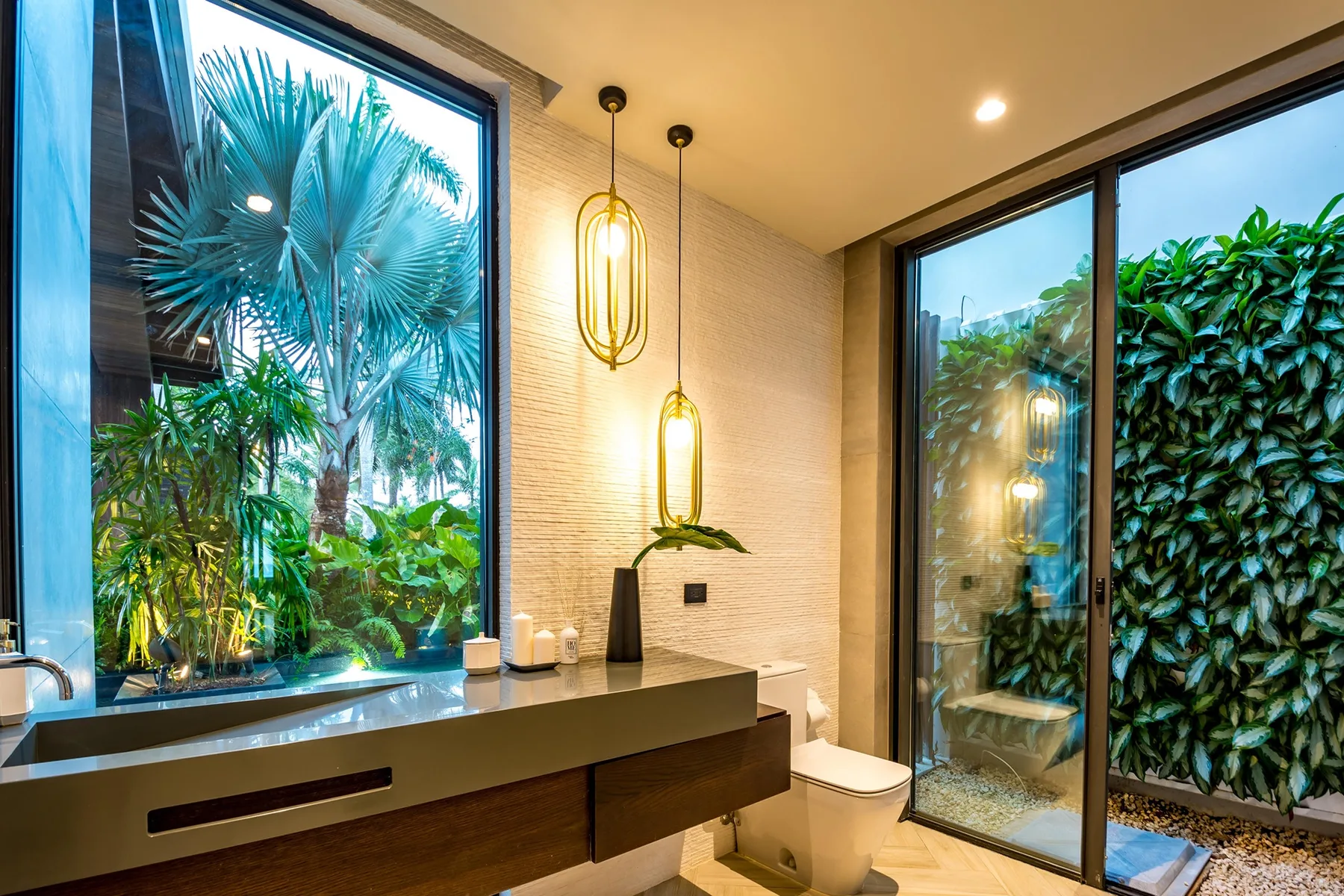 A bathroom vanity and toilet sit beside large windows overlooking lush greenery.