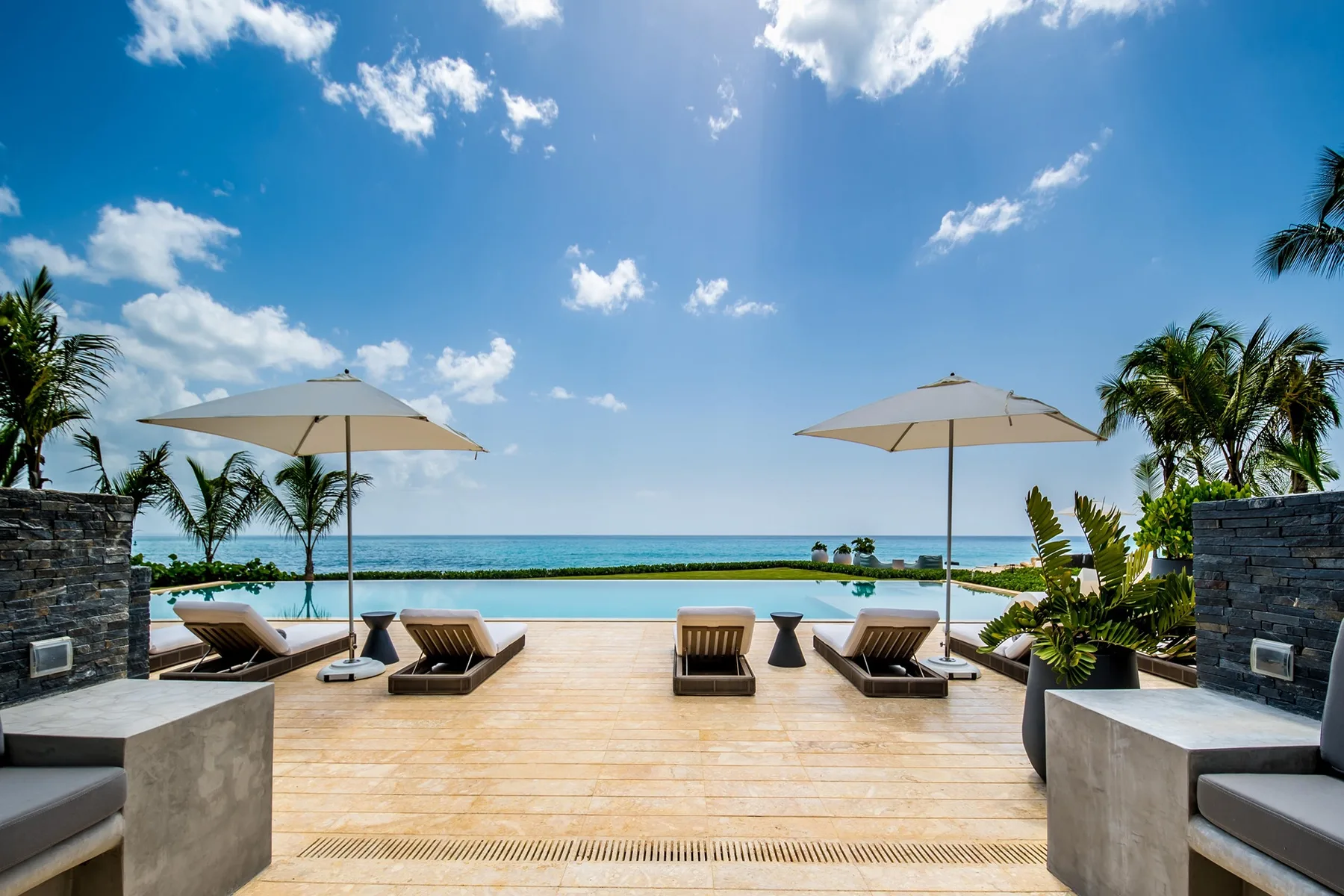A sunny poolside terrace overlooks the ocean, with lounge seating and umbrellas.