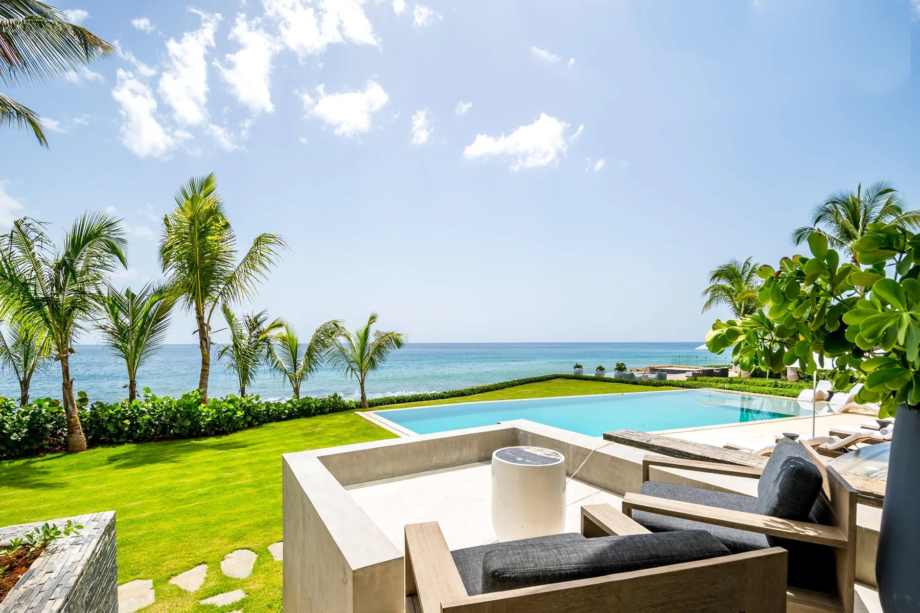 An outdoor poolside seating area overlooks a green lawn and the ocean.