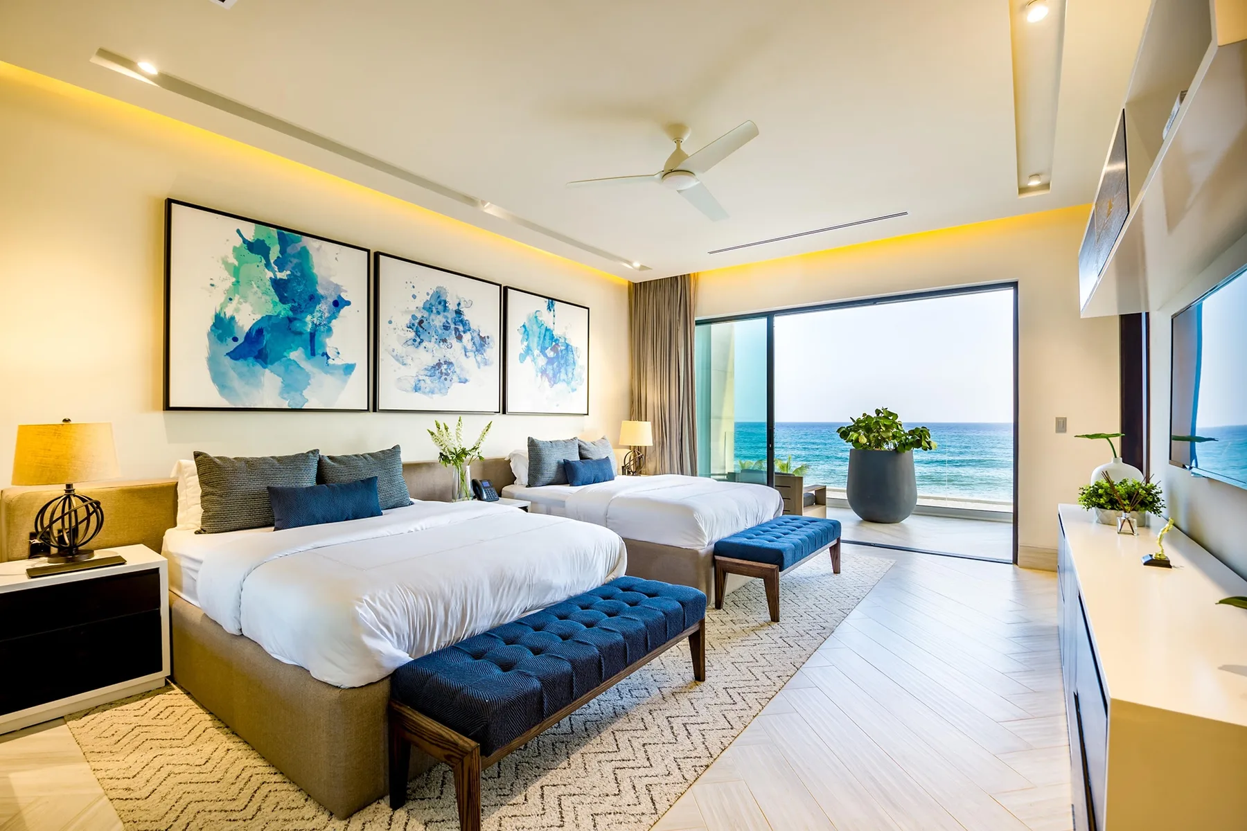 A bright bedroom with two beds, a seating bench, and ocean views through wide windows.