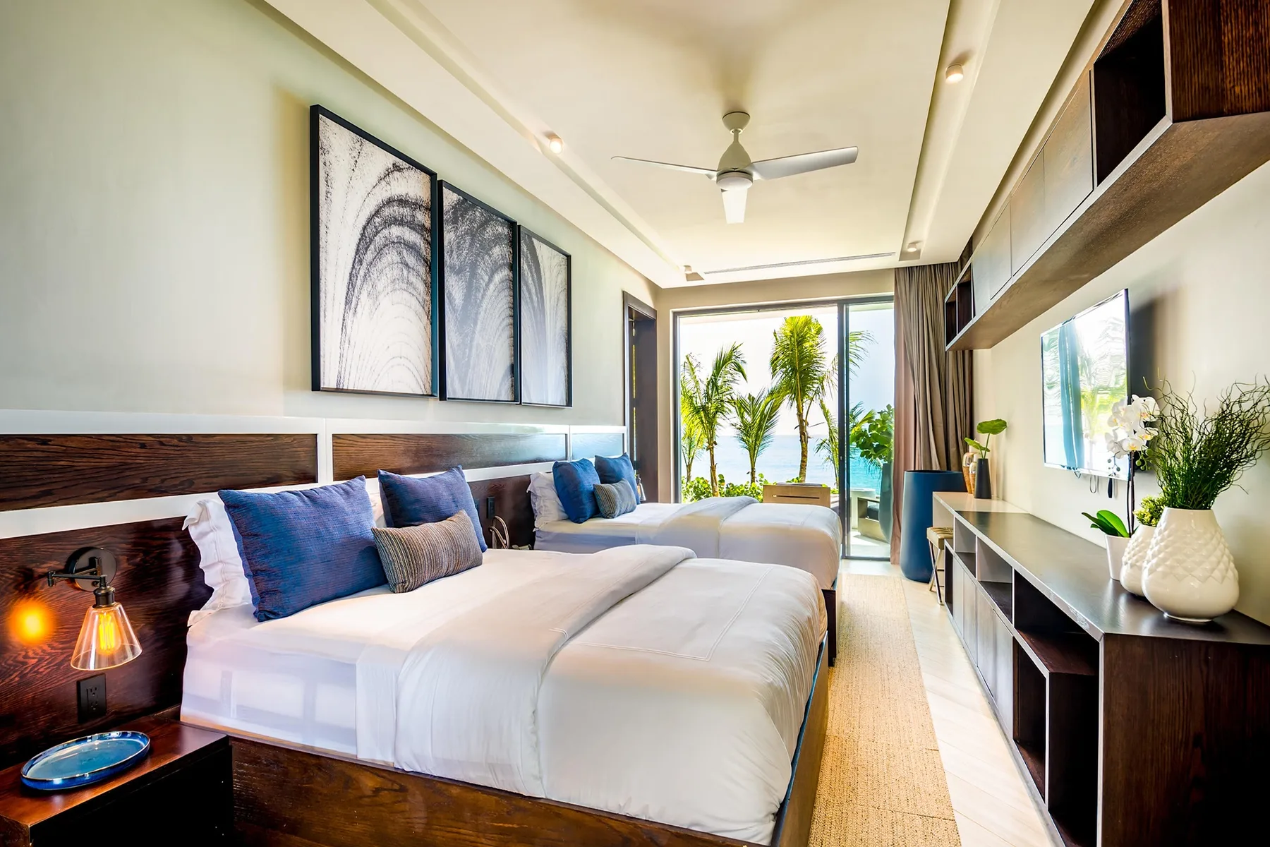 A bright bedroom with two beds, framed artwork, and a ceiling fan.
