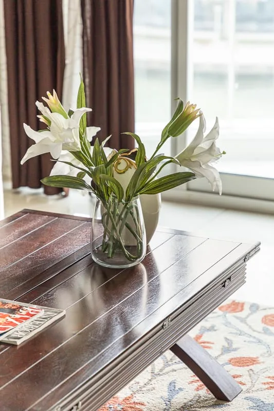 Un jarrón con flores blancas está sobre una mesa de centro de madera junto a una ventana.