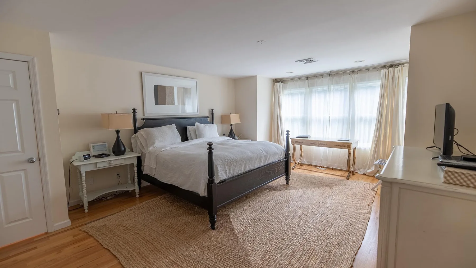 Un dormitorio luminoso con una cama grande y una ventana con cortinas transparentes.