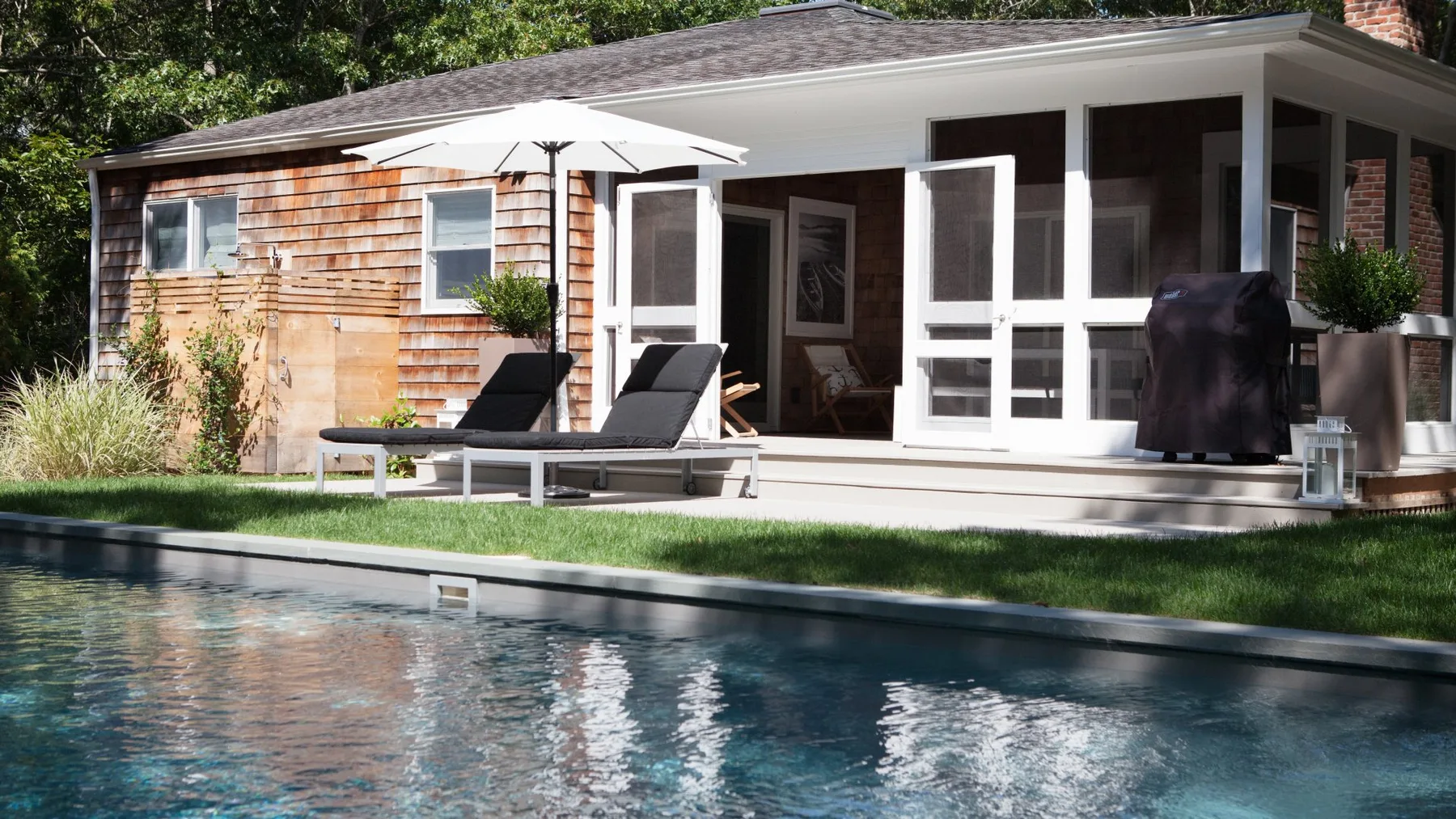 Una piscina junto a un patio con asientos reclinables y una sombrilla blanca.