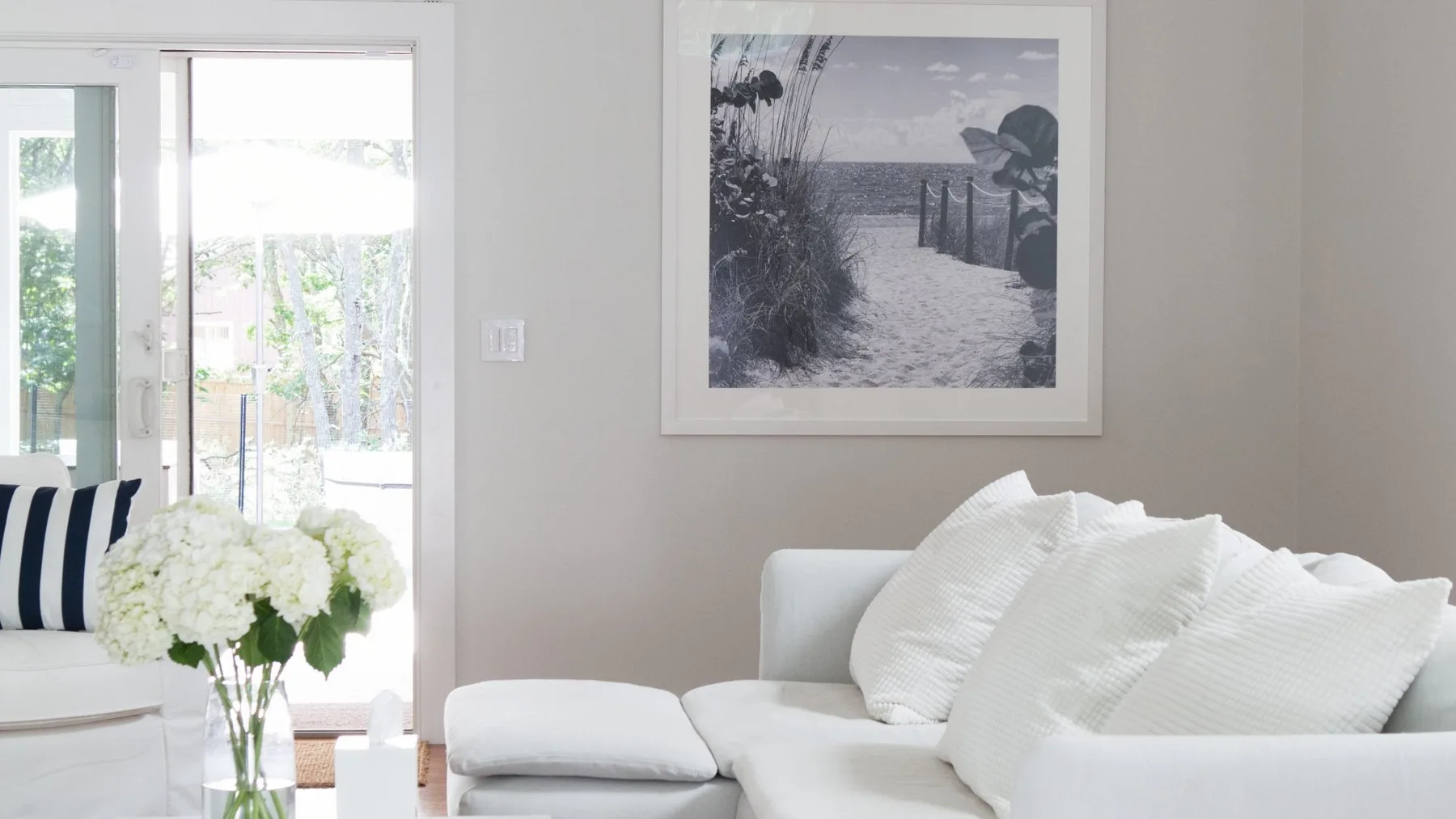 Una sala de estar luminosa con asientos blancos, cojines a rayas y un jarrón con flores.