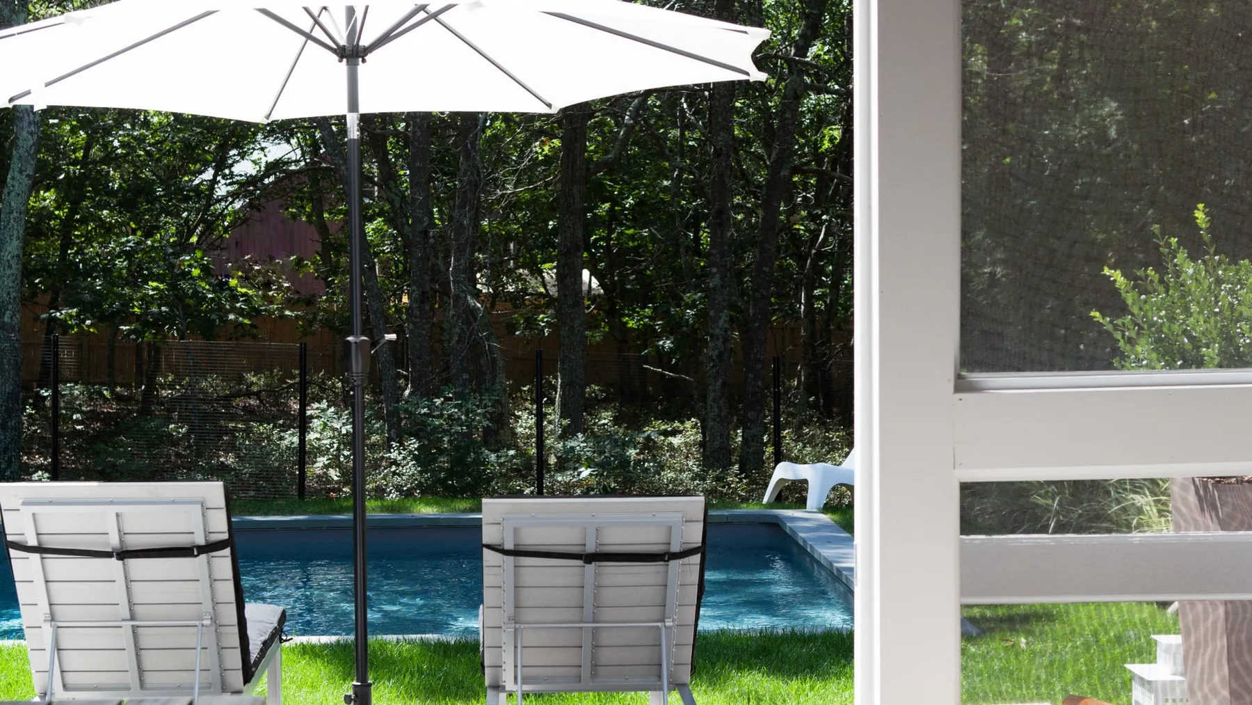 Una piscina exterior con asientos bajo una sombrilla grande, rodeada de árboles.