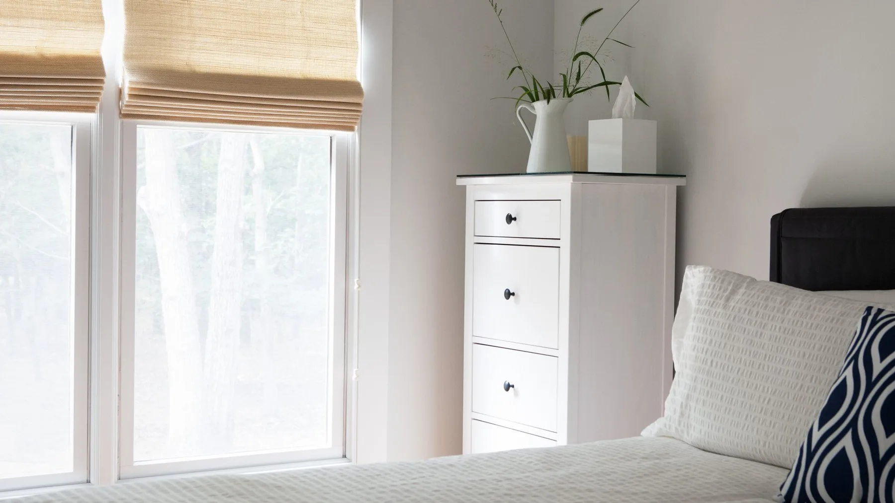 Un dormitorio luminoso con una cama junto a una cómoda blanca y grandes ventanas.