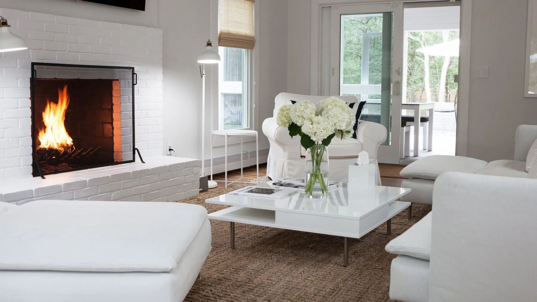 Una sala luminosa con chimenea, asientos blancos y flores sobre una mesa de centro de vidrio.